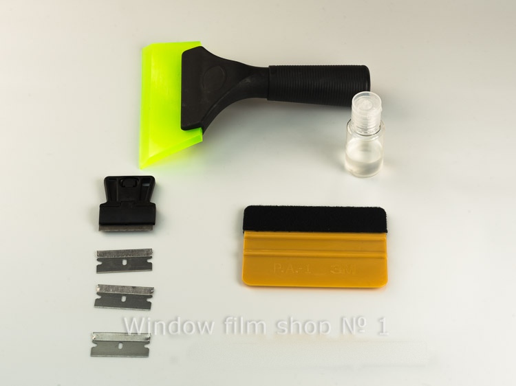 Window tint tool kit laid out: scraper with a green guard, spare blades, yellow squeegee and a bottle of mounting concentrate